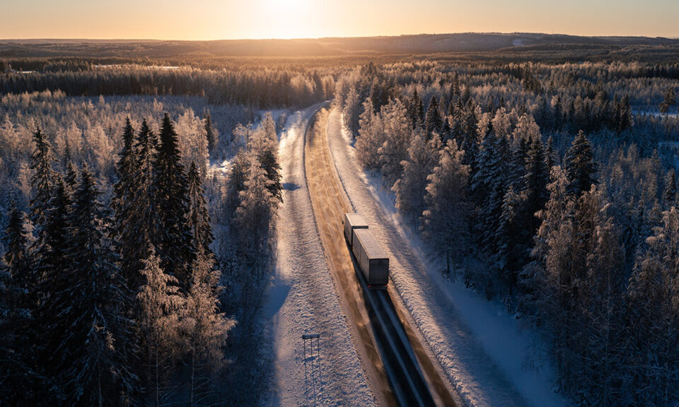 En lastbil kör på en snöklädd väg som är omgärdad av skog. Solen håller på att gå ner. Det är vackert vintrigt.