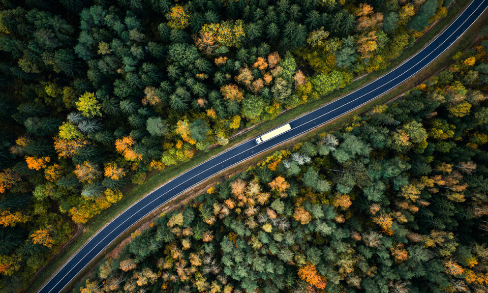 Flygbild på en väg, som går genom en blandskog på hösten, och som en lastbil kör på.
